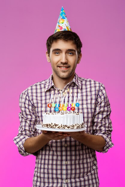 Hombre hermoso joven que sonríe, sosteniendo la torta de cumpleaños sobre la pared púrpura.