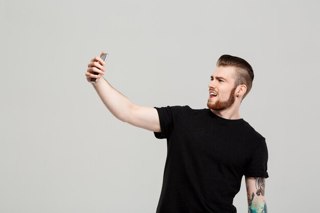 Hombre hermoso joven que sostiene el teléfono que hace el selfie sobre la pared gris.