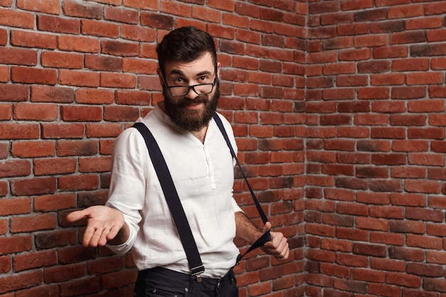 Hombre hermoso joven en vidrios que gesticula en la pared de ladrillo.