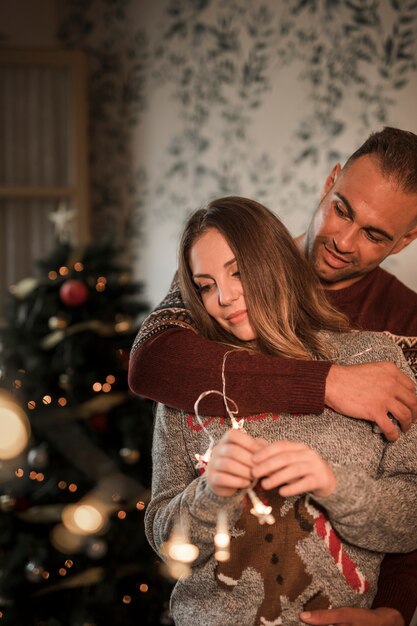 El hombre hermoso que abraza a la mujer alegre en suéteres acerca al árbol de navidad