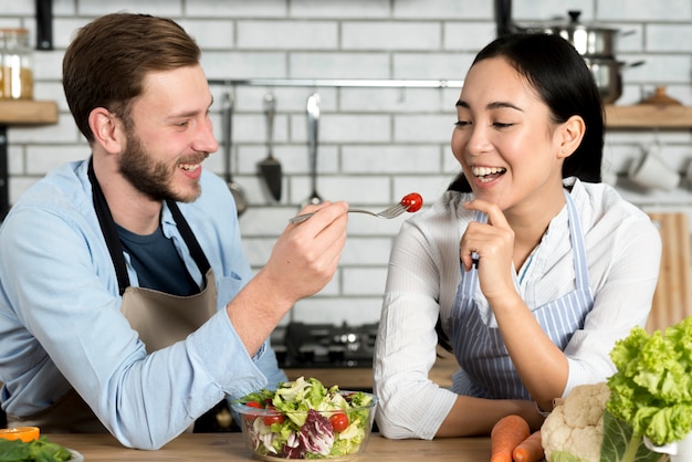 Hombre hermoso que alimenta el tomate alegre a su esposa en cocina