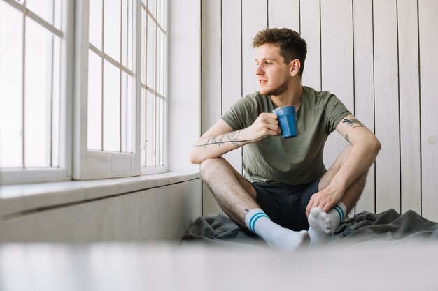 Hombre hermoso que mira a través de la ventana que sostiene la taza de café en la mañana