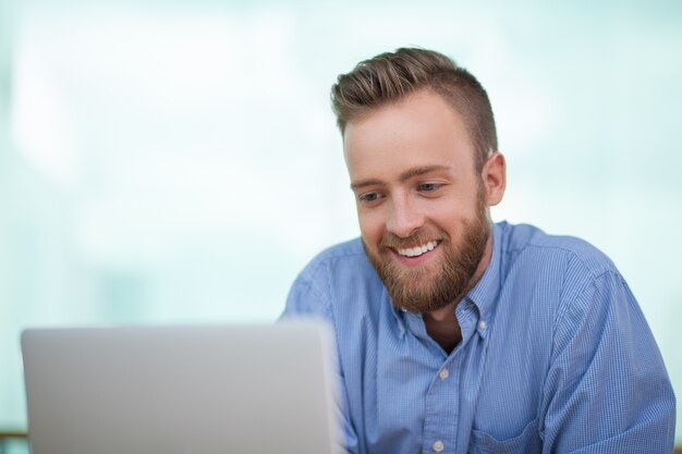 El hombre hermoso sonriente usando la computadora portátil en interiores