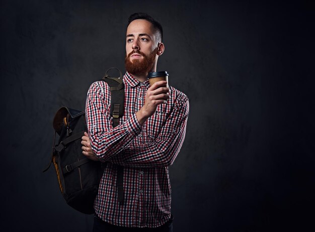 Hombre hipster con barba pelirroja en una camisa de lana roja sostiene el teléfono inteligente sobre fondo gris.