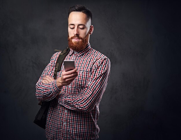 Hombre hipster con barba pelirroja en una camisa de lana roja sostiene el teléfono inteligente sobre fondo gris.