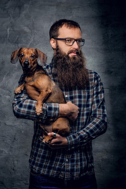 Hombre hipster barbudo con anteojos vestido con una camisa a cuadros sostiene en brazos un lindo perro pequeño.