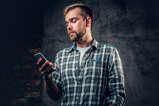 Hombre hipster barbudo en una camisa de lana sostiene un teléfono inteligente sobre fondo gris.
