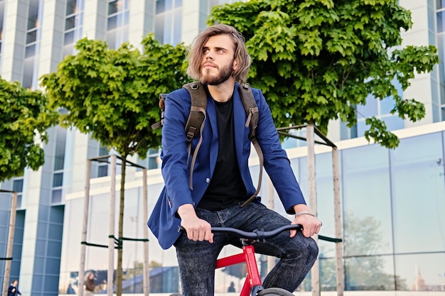 Hombre hipster barbudo con mochila se sienta en la bicicleta roja fija en un parque.