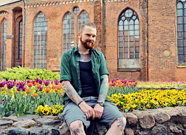 Hombre hipster barbudo tatuado se sienta en un muro de piedra sobre la antigua iglesia en el fondo.