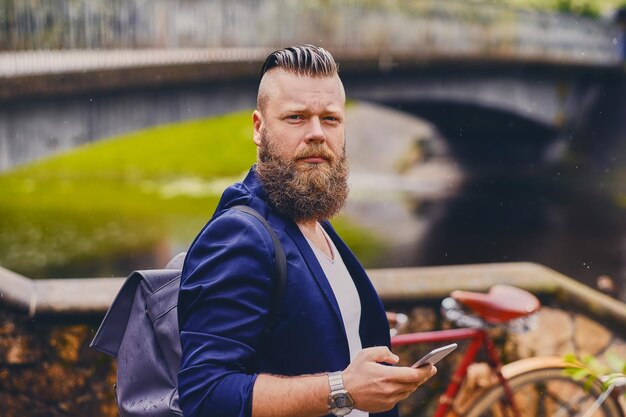 Hombre hipster barbudo usando un teléfono inteligente en un parque cerca del río.