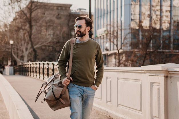 Hombre hipster con estilo atractivo caminando en las calles de la ciudad con bolso de cuero con sudadera y gafas de sol, tendencia de estilo urbano, día soleado