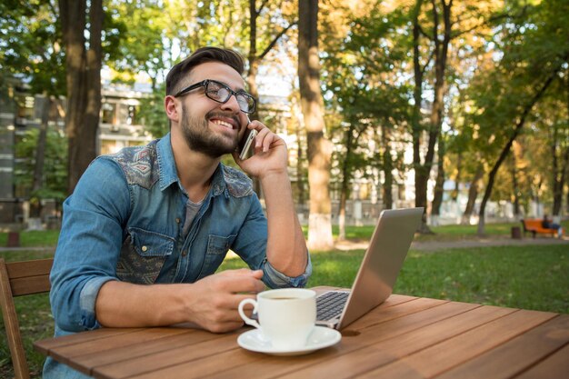 Hombre hipster freelance feliz hablando por teléfono móvil en la mesa de madera Hombre sonriente usando una computadora portátil y tomando café