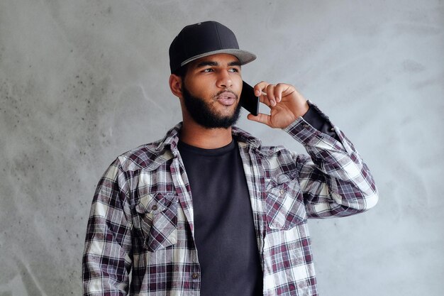 Un hombre hipster negro vestido con una camisa de lana y béisbol se sienta cerca de la ventana y usa un teléfono inteligente.