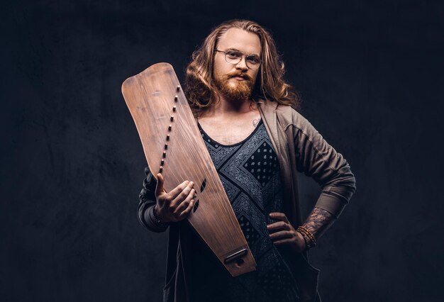 Hombre hipster pelirrojo con cabello largo y exuberante y barba llena vestido con ropa informal sostiene un instrumento musical tradicional ruso - gusli. Aislado en un fondo oscuro.