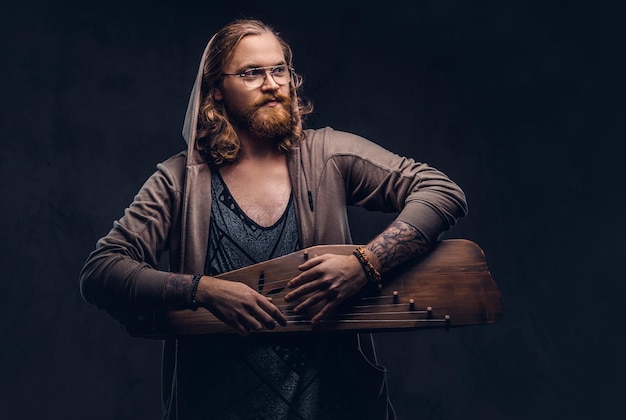 Hombre hipster pelirrojo con cabello largo y exuberante y barba llena vestido con una sudadera con capucha y una camiseta tocando un instrumento musical tradicional ruso - gusli. Aislado en el fondo oscuro.