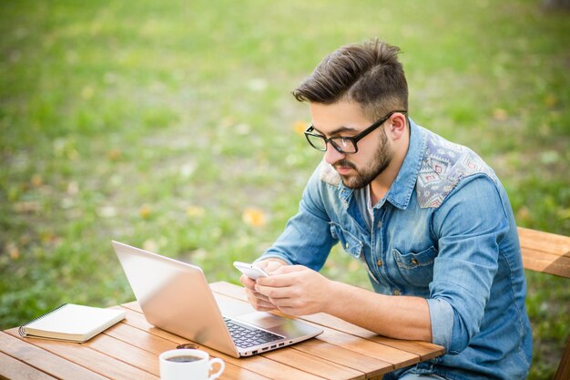 Hombre hipster serio freelance usando su teléfono inteligente Hombre guapo trabajando con su computadora en el parque