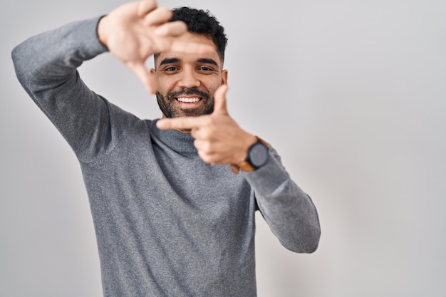 Hombre hispano con barba de pie sobre fondo blanco sonriendo haciendo marco con manos y dedos con cara feliz creatividad y concepto de fotografía