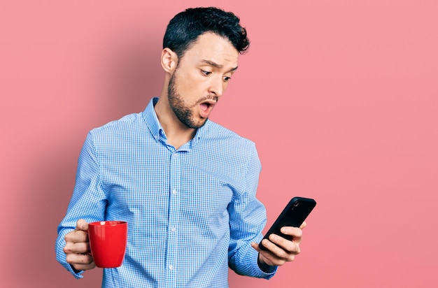 Foto gratuita hombre hispano con barba usando un teléfono inteligente y bebiendo una taza de café con cara de shock mirando escéptico y sarcástico sorprendido con la boca abierta