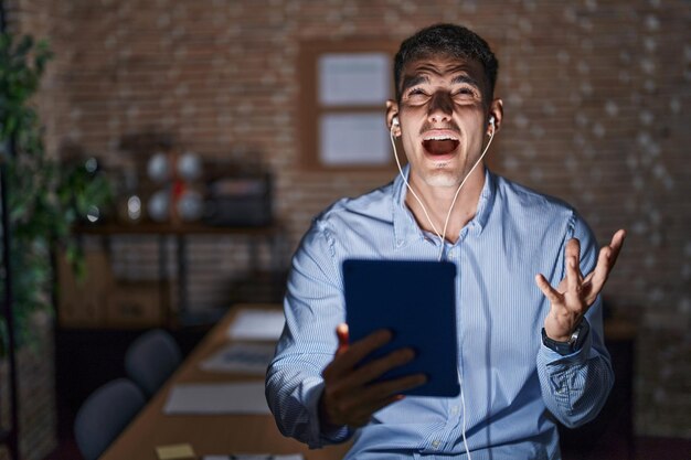 Hombre hispano guapo trabajando en la oficina por la noche loco y enojado gritando y gritando con expresión agresiva y brazos levantados concepto de frustración