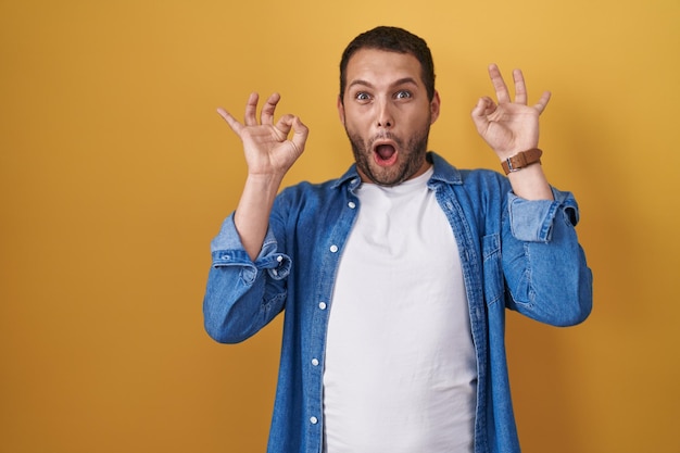 Foto gratuita hombre hispano parado sobre un fondo amarillo que parece sorprendido y conmocionado haciendo un buen símbolo de aprobación con los dedos. expresión loca