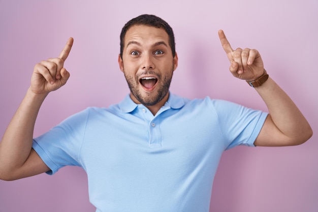 Hombre hispano de pie sobre fondo rosa sonriendo asombrado y sorprendido y señalando con los dedos y los brazos levantados