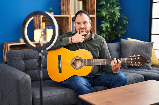 Hombre hispano de talla grande con barba tocando la guitarra clásica en el estudio de música grabándose a sí mismo con cara seria pensando en la pregunta con la mano en la barbilla, pensando en una idea confusa