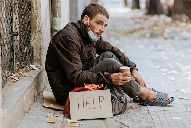 Hombre sin hogar al aire libre con taza y señal de ayuda