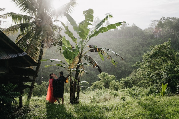 Hombre de impermeable negro tocando la cara de la novia en la naturaleza. Pareja de turistas posando en la selva tropical.