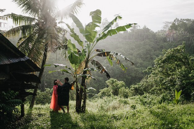 Hombre de impermeable negro tocando la cara de la novia en la naturaleza. Pareja de turistas posando en la selva tropical.