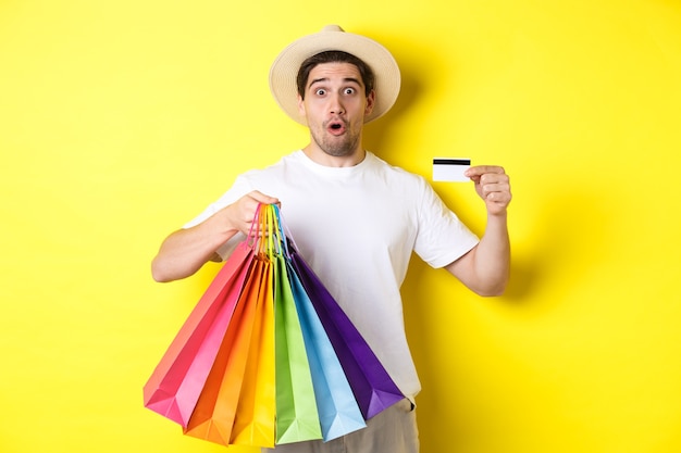 Hombre impresionado mostrando bolsas de la compra con productos y tarjeta de crédito, de pie sobre fondo amarillo.