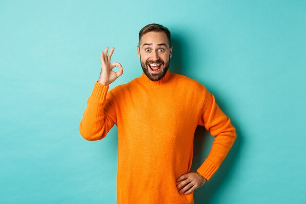 Hombre impresionado recomendando promoción, mostrando el signo de bien y sonriendo asombrado, aprobado y de acuerdo, de pie sobre una pared turquesa claro.