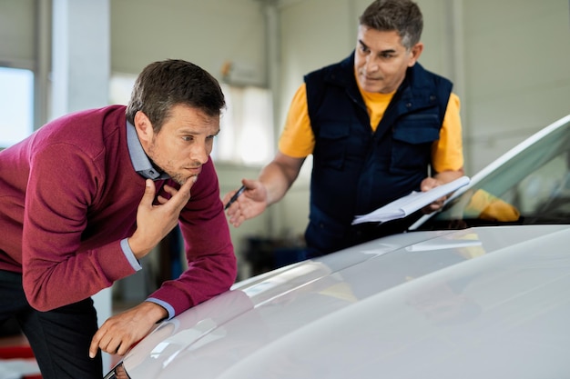 Hombre incierto examinando el capó de su auto mientras el mecánico habla con él en el taller de reparación de automóviles