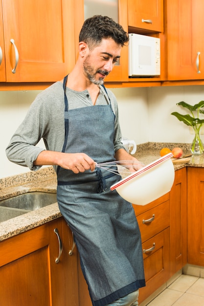 Hombre inclinado cerca del mostrador de la cocina batió el huevo con un batidor en el tazón.