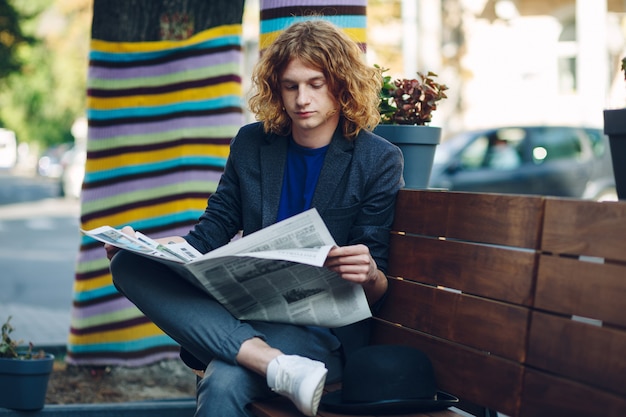 Hombre inconformista de pelo rojo sentado en un banco leyendo un periódico