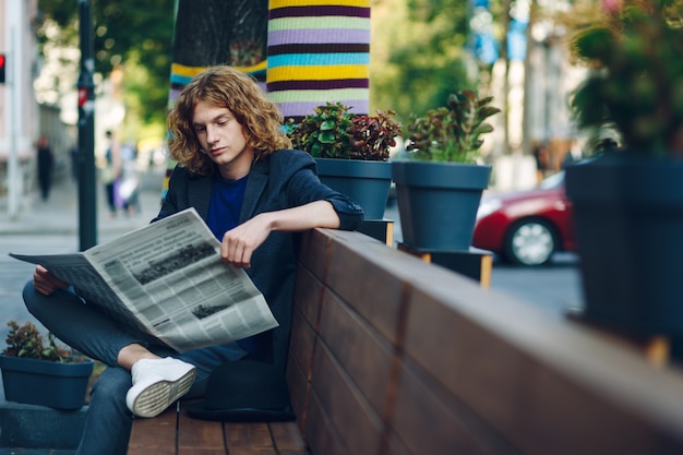 Hombre inconformista de pelo rojo sentado en un banco leyendo un periódico