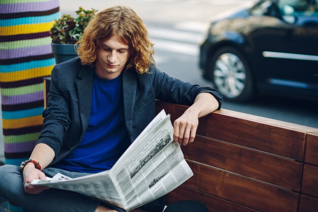 Hombre inconformista de pelo rojo sentado en un banco leyendo un periódico