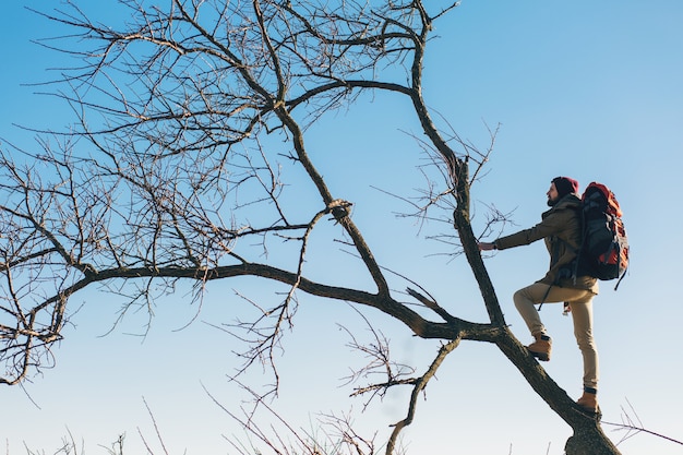 Hombre inconformista viajando con mochila, de pie en un árbol contra el cielo, con chaqueta abrigada, turista activo, explorando la naturaleza en la estación fría