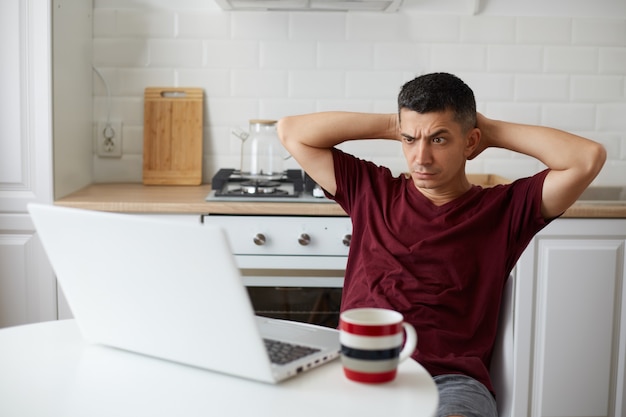 Hombre independiente desconcertado con ropa de estilo casual sentado frente a una computadora portátil en la cocina, mantiene los brazos detrás de la cabeza, mirando la pantalla de la computadora portátil con expresión confusa, con el ceño fruncido.