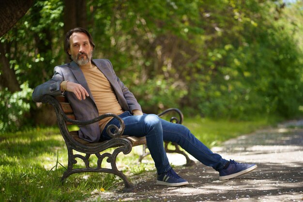 Un hombre independiente maduro con barba gris pasa tiempo sentado en el banco del parque disfrutando del tiempo libre o esperando a sus colegas con chaqueta informal y jeans