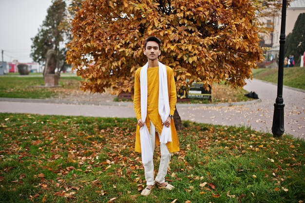 Hombre indio con estilo en ropa tradicional amarilla con bufanda blanca posada al aire libre contra hojas de otoño