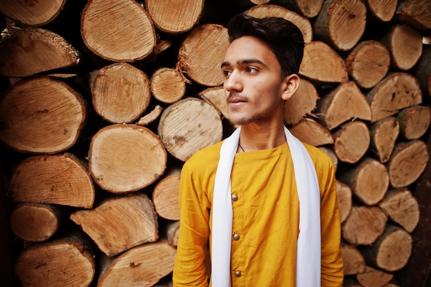 Hombre indio con estilo en ropa tradicional amarilla con bufanda blanca posada al aire libre contra tocones de madera