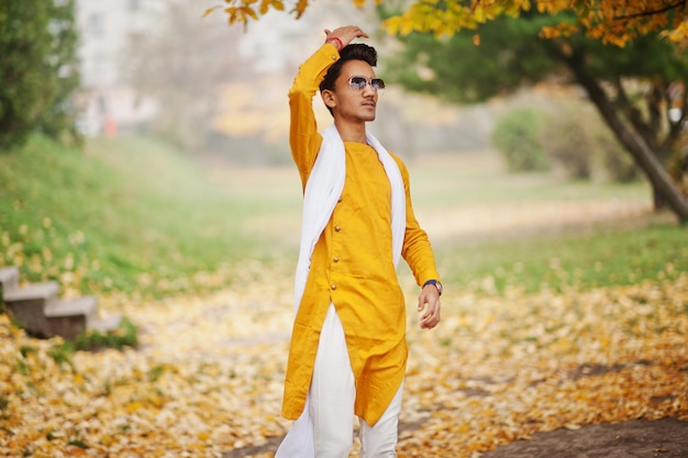 Hombre indio con estilo en ropa tradicional amarilla con gafas de sol de bufanda blanca posadas al aire libre contra el árbol de hojas de otoño