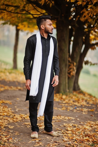 Hombre indio con estilo en ropa tradicional negra con bufanda blanca posada al aire libre contra el árbol de hojas amarillas de otoño