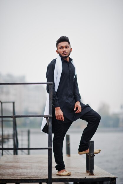 Hombre indio con estilo en ropa tradicional negra con bufanda blanca posada al aire libre contra el muelle de niebla del mar