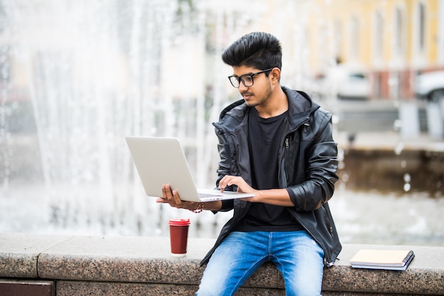 Hombre indio guapo con laptop mientras está sentado cerca de la fuente en el centro de la ciudad en un día