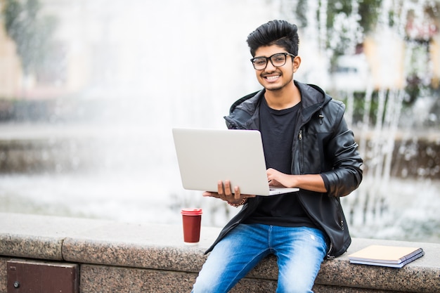 Hombre indio guapo con laptop sentado cerca de la fuente en el centro de la ciudad