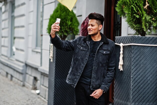 Hombre indio guapo y de moda en chaqueta de jeans negros posó al aire libre y haciendo selfie en el teléfono