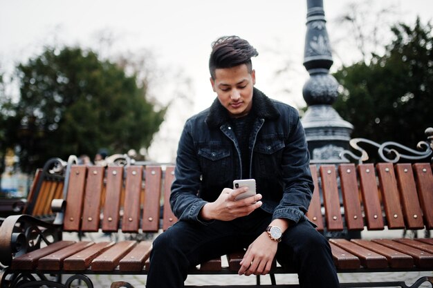 Hombre indio guapo y de moda con chaqueta de jeans negros posó al aire libre sentado en un banco con el teléfono móvil a mano