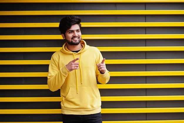Hombre indio joven hipster urbano en una sudadera amarilla de moda Chico fresco del sur de Asia usa sudadera con capucha contra un fondo rayado mostrando los dedos para liberar espacio