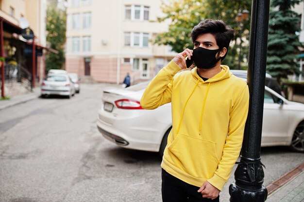 Hombre indio joven hipster urbano en una sudadera amarilla de moda con teléfono móvil Chico fresco del sur de Asia usa sudadera con capucha y máscara protectora de cara negra durante la nueva normalidad